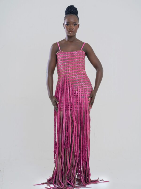 La robe Terra Rouge est une œuvre en devenir, une construction en cours, un hommage aux bâtisseuses de leur propre destin. Inspirée des maisons en terre rouge des peuples Bamiléké et d’autres ethnies africaines, elle traduit l’idée d’un édifice qui prend forme, d’un avenir qui se façonne à chaque instant. Son tissage en bordeaux profond et fuchsia doux évoque les briques de terre rouge, ces murs chargés d’histoire et de mémoire. Chaque fil entrelacé rappelle les mains des artisans bâtissant, pierre après pierre, un abri, un foyer, une identité. La silhouette droite, d’une élégance épurée, descend jusqu’au genou avant de laisser place à une cascade de franges en fils tissés, représentant un toit de paille en cours d’installation. À chaque mouvement, ces fils dansent comme les herbes séchées sous le vent, donnant vie à cette pièce unique. Les fines bretelles, délicates et discrètes, apportent une touche de légèreté et de modernité, contrastant avec la puissance du tissage et la symbolique architecturale de la robe. Porter la robe Terre Rouge, c’est affirmer sa force et son ancrage tout en embrassant la fluidité du changement. Une maison en construction, un chemin en devenir, une histoire à écrire.